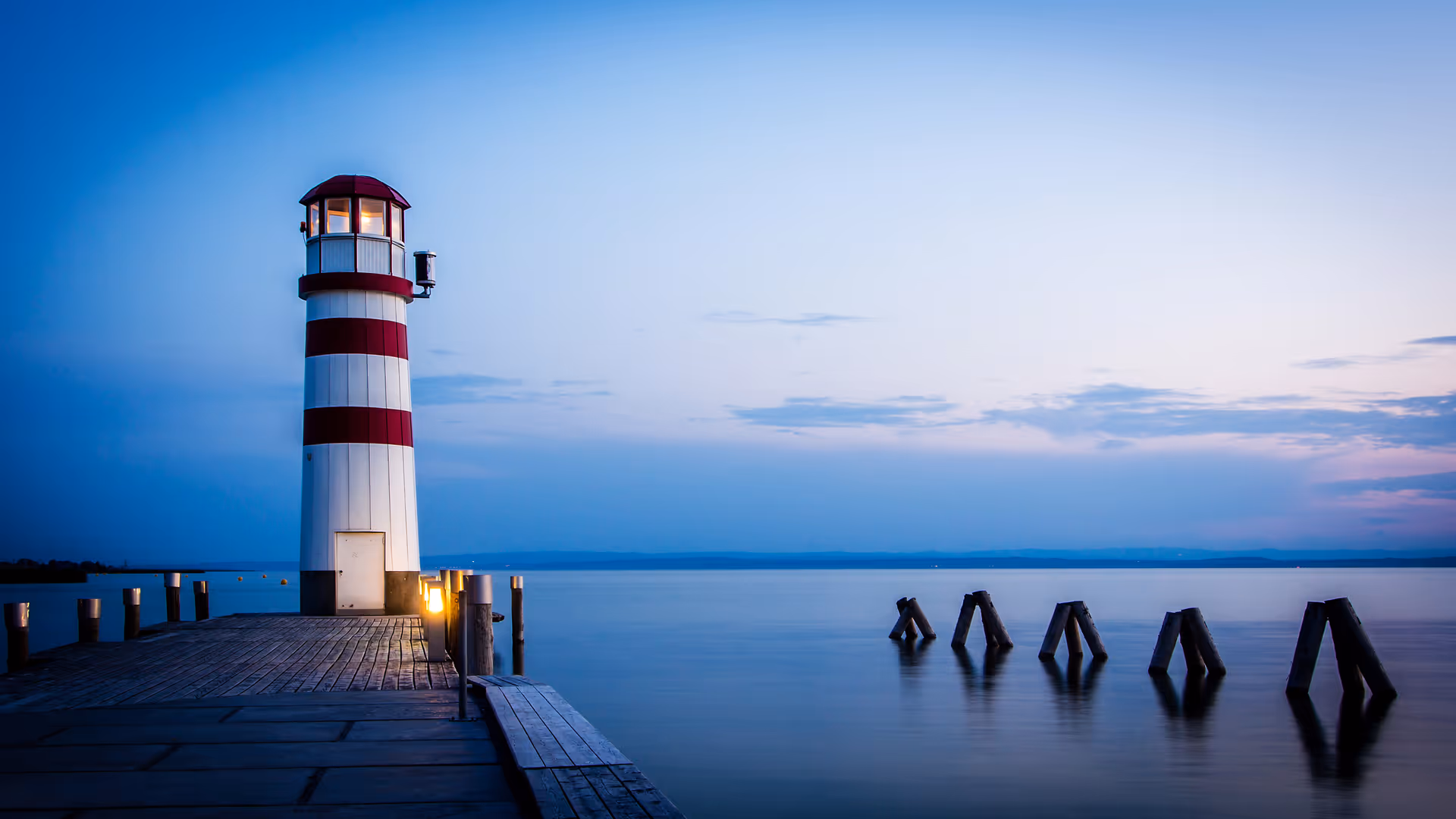 A white lighthouse with red stripes stands at the end of a wooden pier over water.