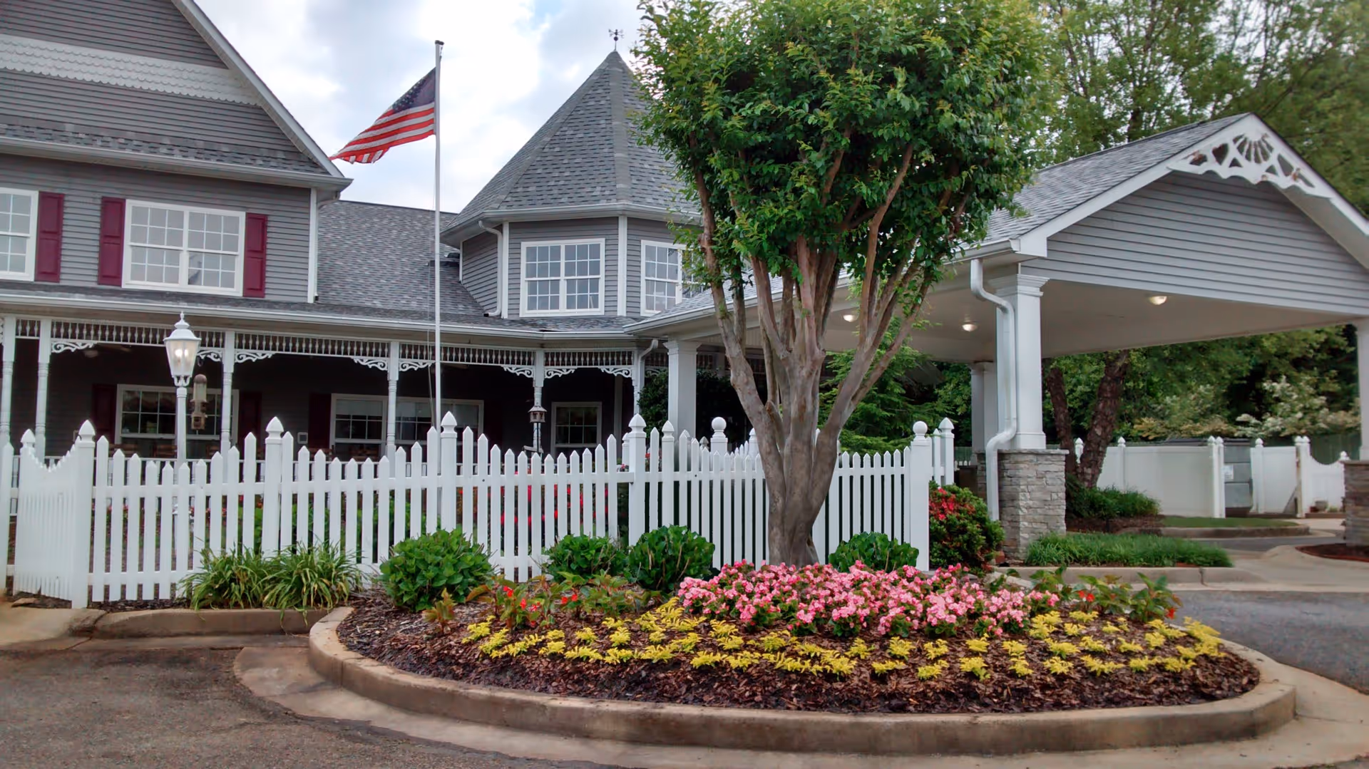 Exterior view of a senior living facility named Country Village Senior Living featuring a gray building with white trim, a white picket fence, a landscaped flower bed with pink and yellow flowers, a tree, and an American flag on a flagpole.