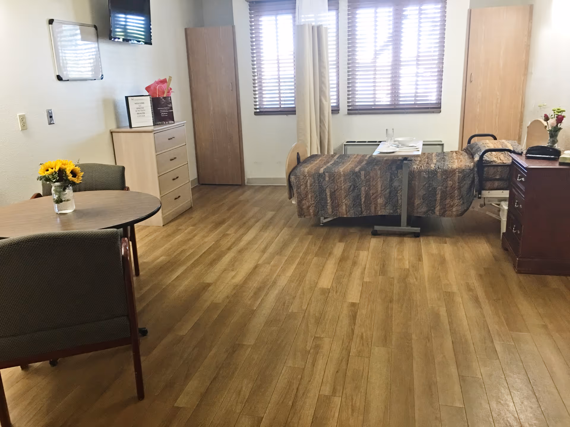 A furnished interior patient room with a bed, round table, dresser, and sunflowers.
