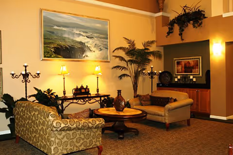 A cozy living room area with two patterned sofas facing each other around a round wooden coffee table. The room features warm yellow walls, a large framed painting of a coastal scene, two table lamps on a console table, decorative plants, and wall sconces providing soft lighting.