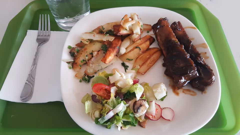 A white plate on a green tray containing two ribs with barbecue sauce, seasoned potato wedges, and a mixed salad with lettuce, tomato, mushrooms, and radishes. A fork on a white napkin and a glass of water are placed next to the plate.