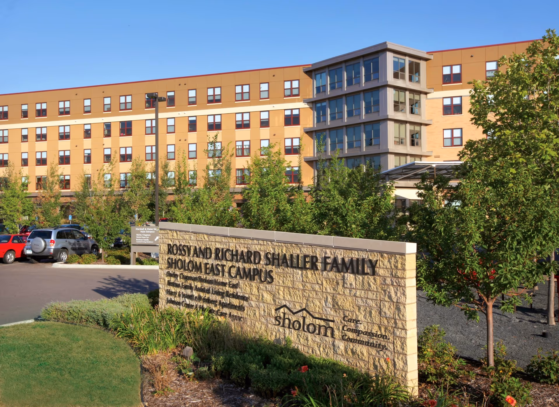 Front exterior of the Rossy and Richard Shaller Family Sholom East Campus building with a stone entrance sign, landscaping, and parked cars.