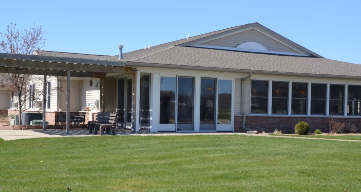 Single-story senior living building with a covered patio, benches and large windows facing a grassy lawn.