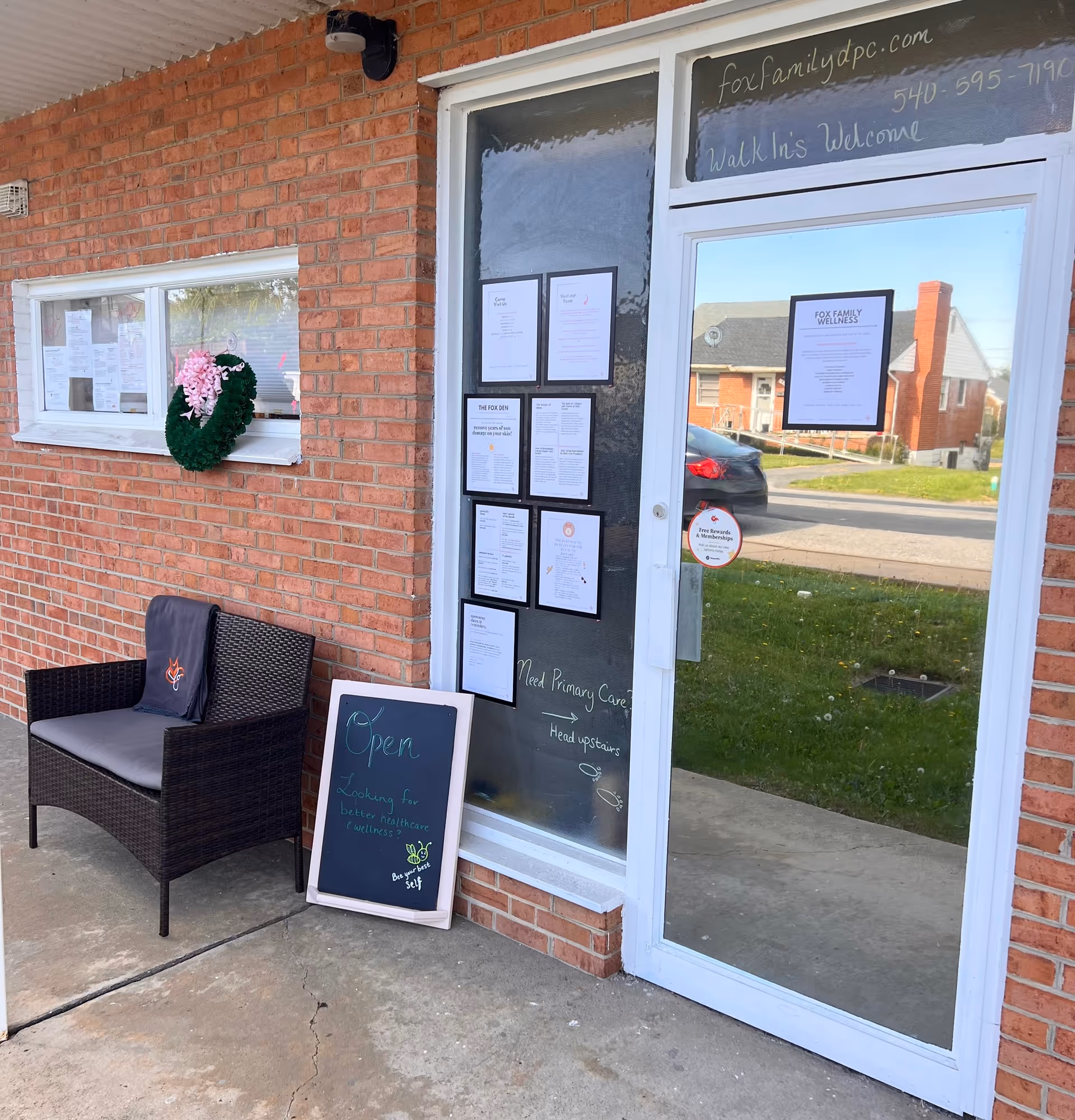 Brick building entrance with glass door, notice-filled window, chalkboard sign, and wicker chair.