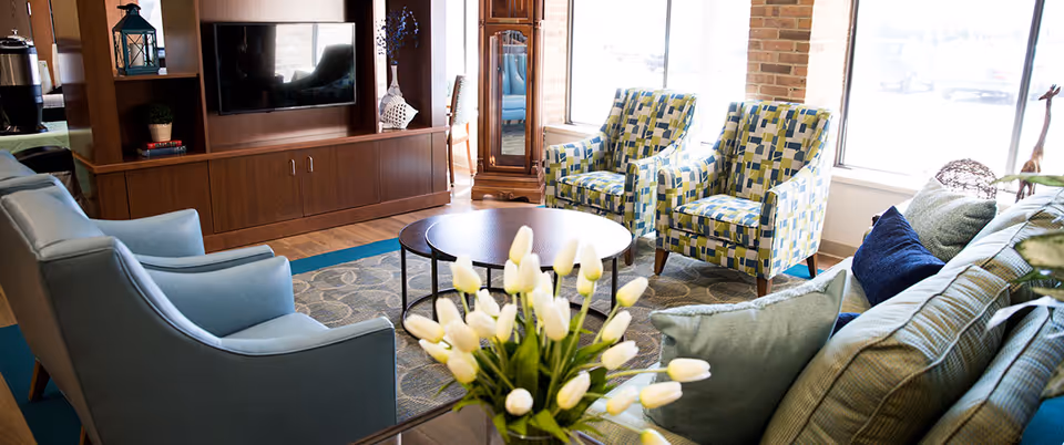 Bright lounge area with sofas, two patterned armchairs, a round coffee table, a TV cabinet, and a vase of white tulips.