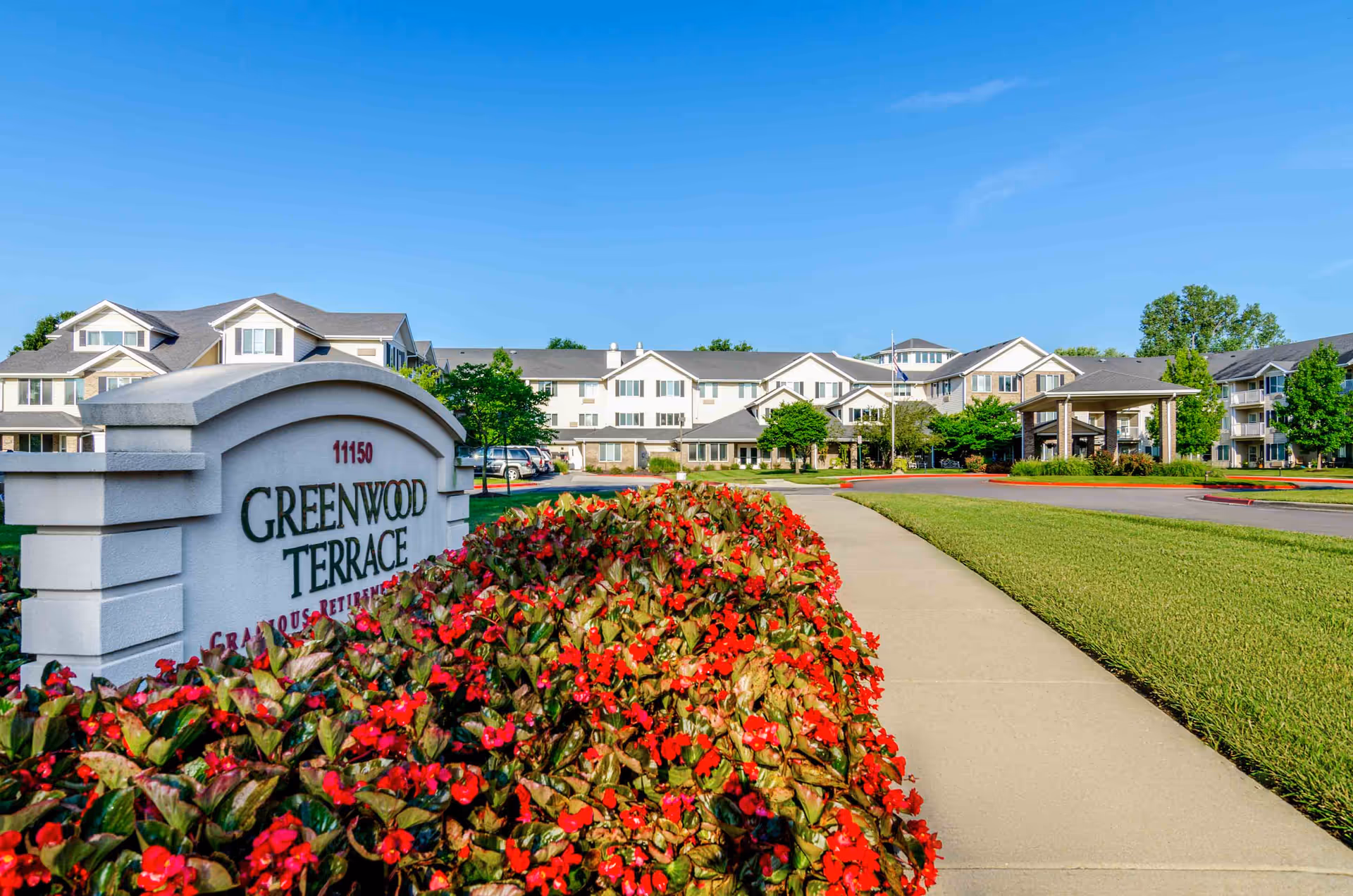 Entrance sign reading "Greenwood Terrace" beside red flowers, a sidewalk, and the senior living building in the background under a blue sky.