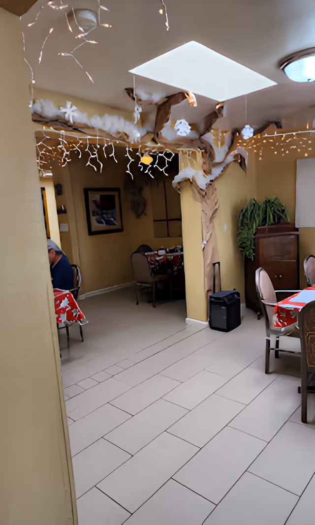 Interior dining area with tiled floor, tables covered in red tablecloths, chairs, decorative ceiling beams and string lights.