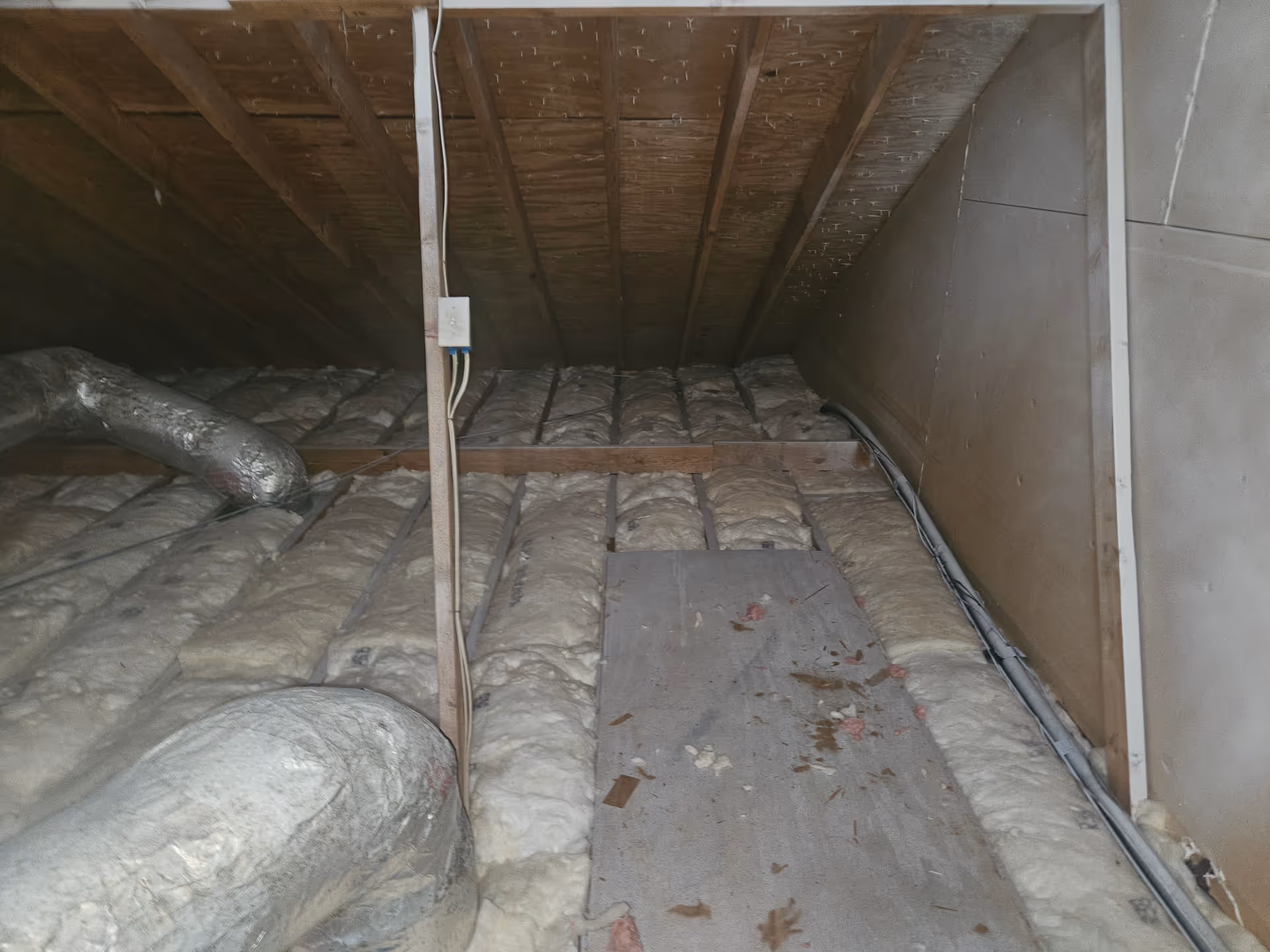 Dusty attic crawl space with exposed insulation, wooden joists, pipes, and debris
