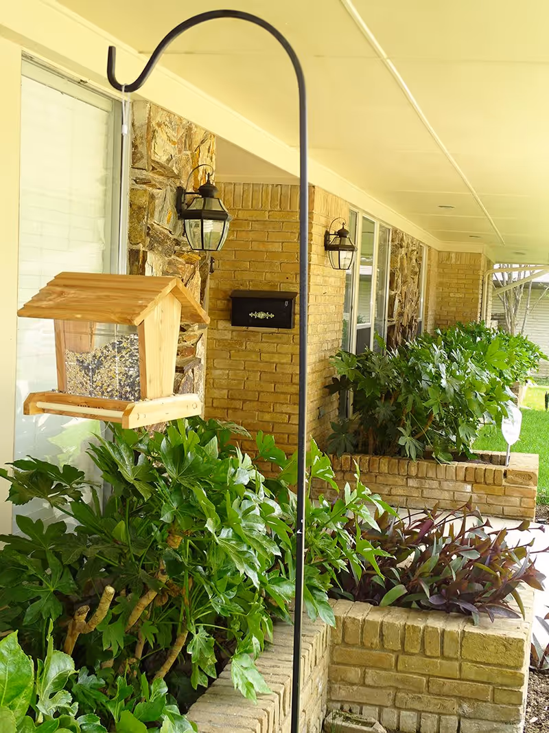 Covered front porch with a hanging wooden bird feeder, brick planters full of green foliage, wall lanterns, and a mailbox.
