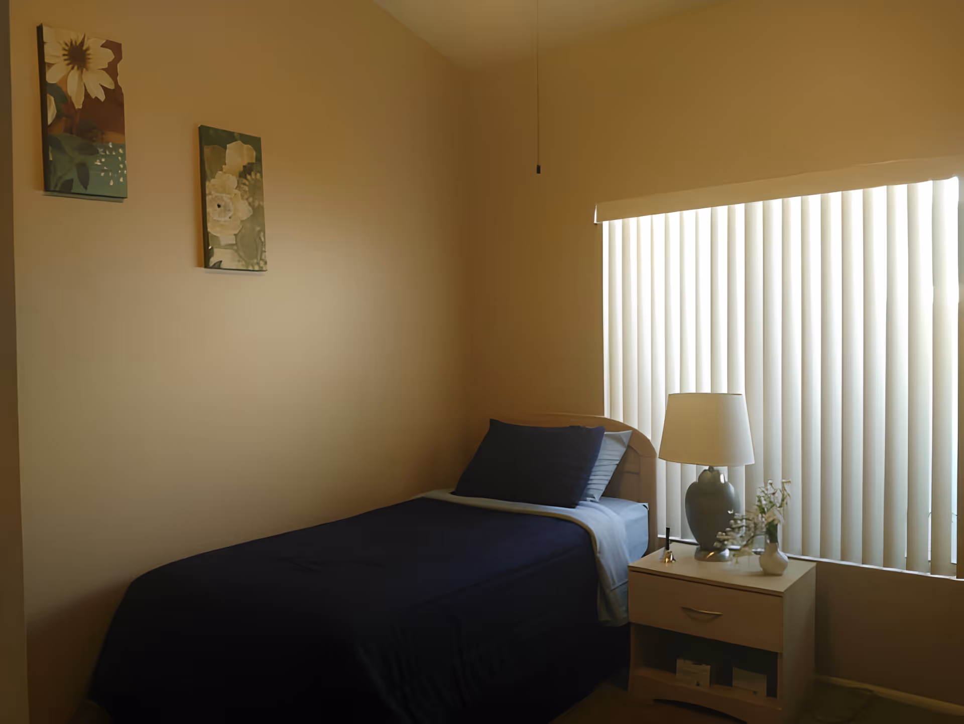 Small single-occupancy bedroom with a bed, nightstand holding a lamp and vase, and a window with vertical blinds.