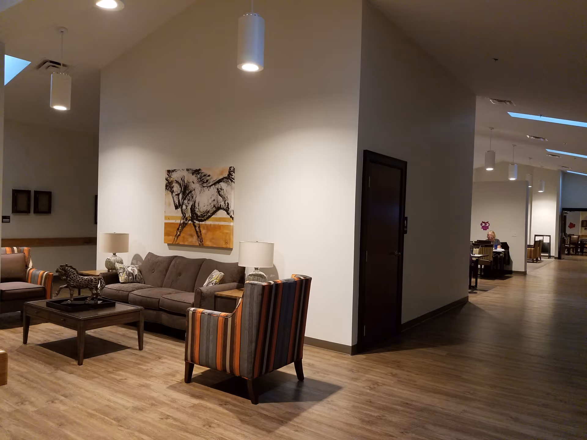 Communal seating area with a sofa, striped armchairs, coffee table, lamps, and wall art in a senior living facility hallway.