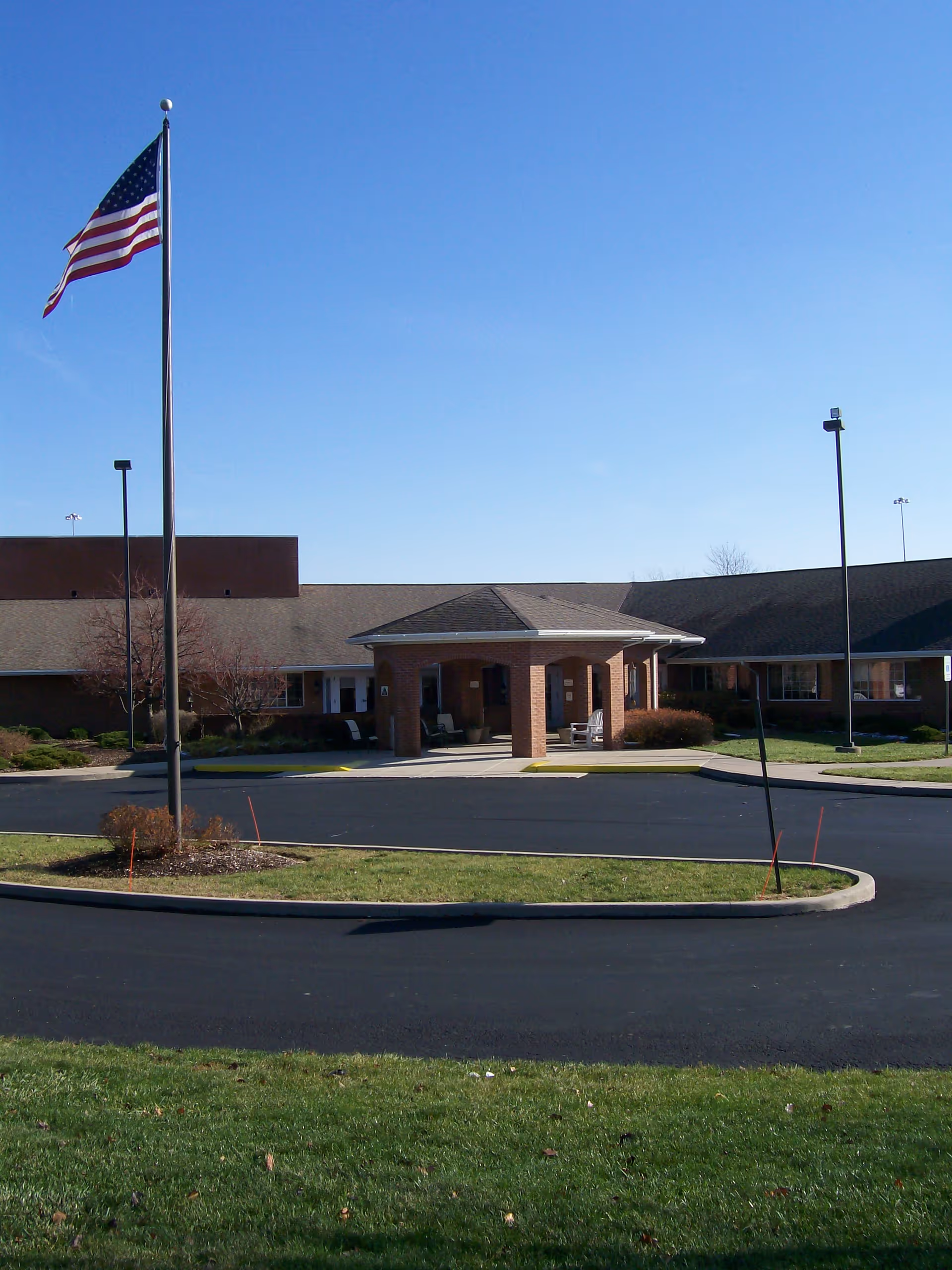 Front exterior of a single-story brick senior living facility with a covered entrance, flagpole, driveway, and lawn.