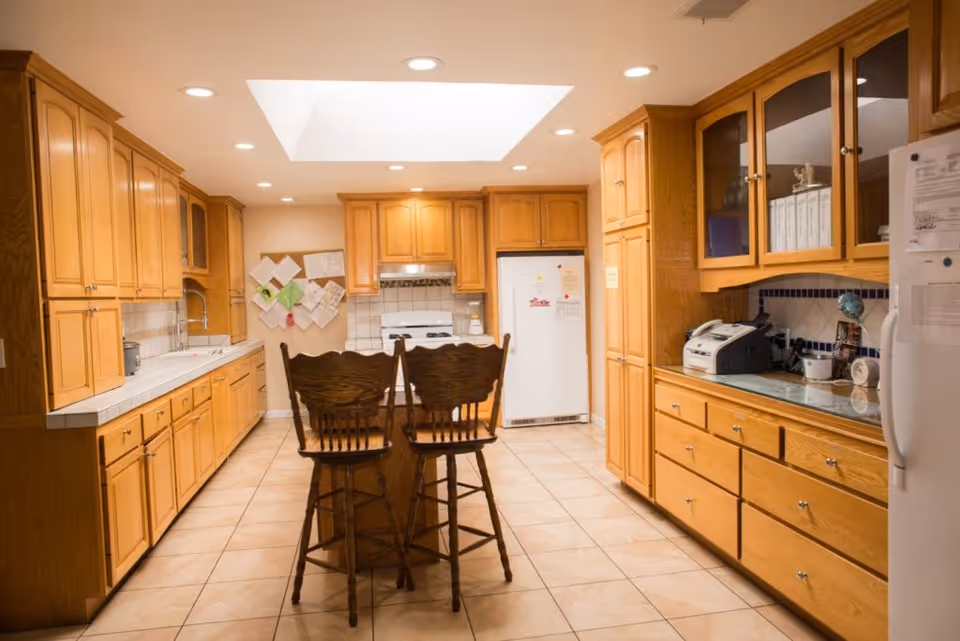 A spacious kitchen with wooden cabinets and drawers on both sides, a white refrigerator, a white stove, and a kitchen island with two wooden chairs. The floor is tiled, and there is a skylight in the ceiling providing natural light. Various papers and notes are pinned on a board on the far wall.