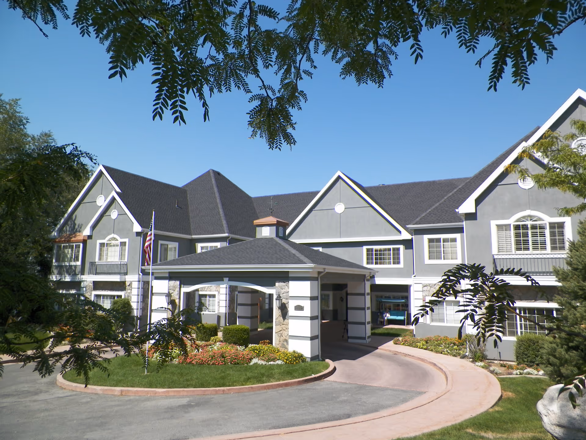 Front exterior of a two-story senior living building with a covered porte-cochere, circular driveway, landscaping, and an American flag.