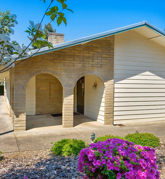 Exterior view of a building with beige brick arches and light-colored siding under a clear blue sky. In the foreground, there are green shrubs and vibrant purple flowers in a landscaped garden area.