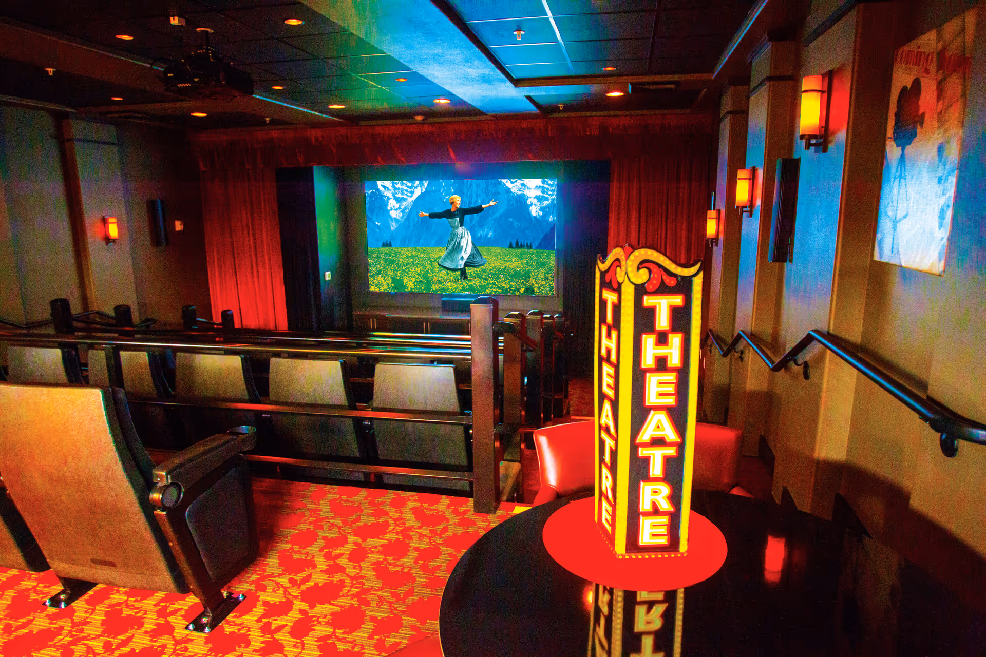 Interior view of a small theater room with rows of cushioned seats facing a large screen showing a scene from a movie with a woman dancing in a field with mountains in the background. The room has red patterned carpet, wall sconces with warm lighting, and a decorative illuminated sign that reads 'THEATRE'.