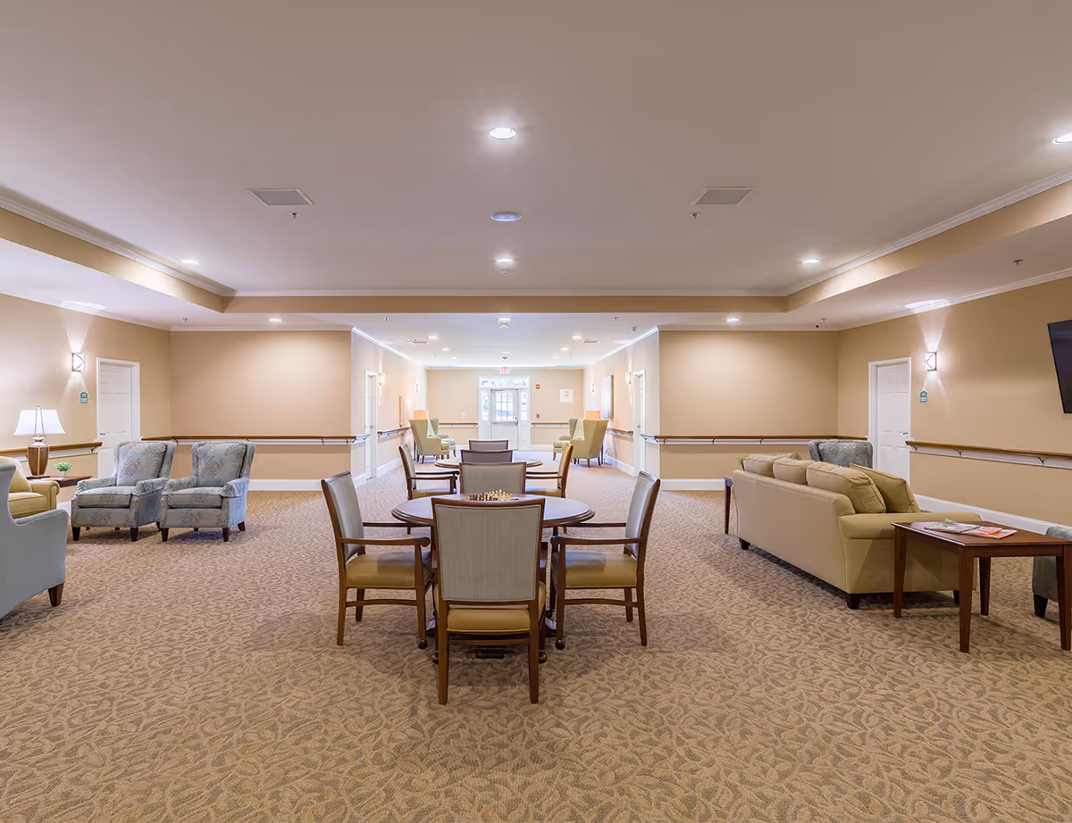 A spacious senior living lounge with beige walls, carpet, sofas, and round tables.