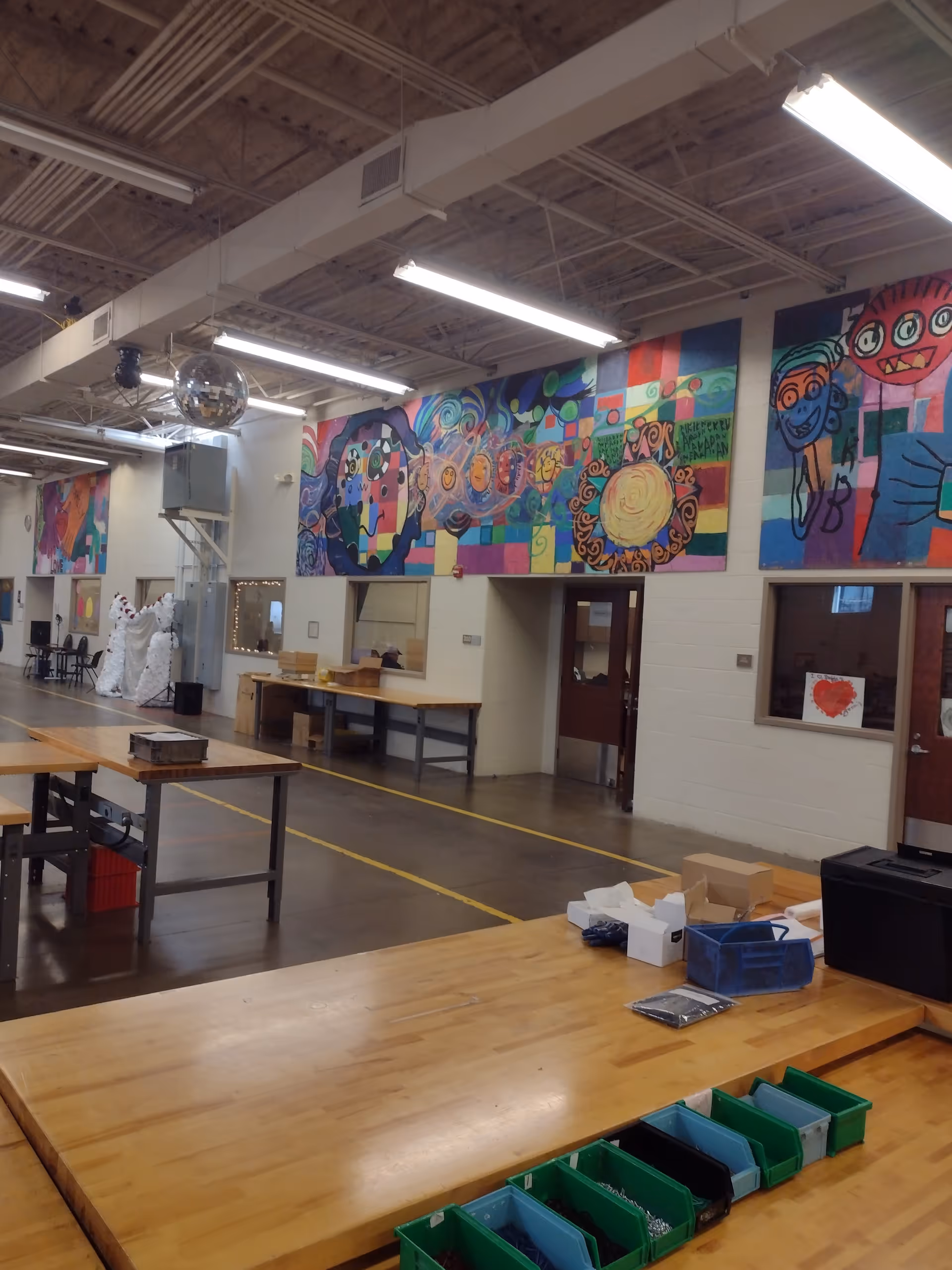 Interior activity room with colorful murals, long tables, and storage bins arranged along the floor.