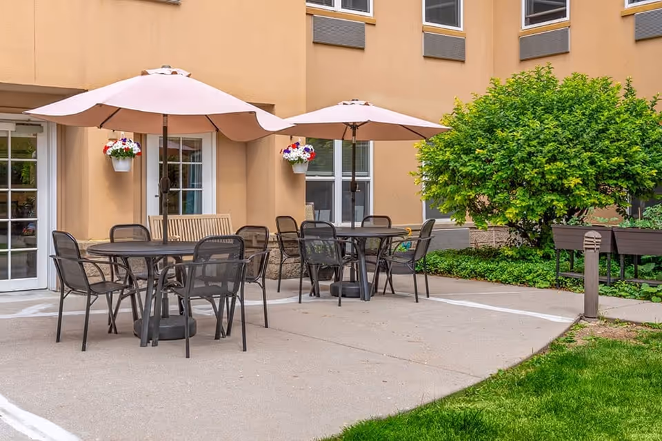 Outdoor patio area with two round tables, each surrounded by black metal chairs and shaded by large beige umbrellas. The patio is adjacent to a beige building with windows and a door. There are hanging flower pots with red, white, and purple flowers on the wall, a wooden bench, green shrubs, and a grassy area nearby.