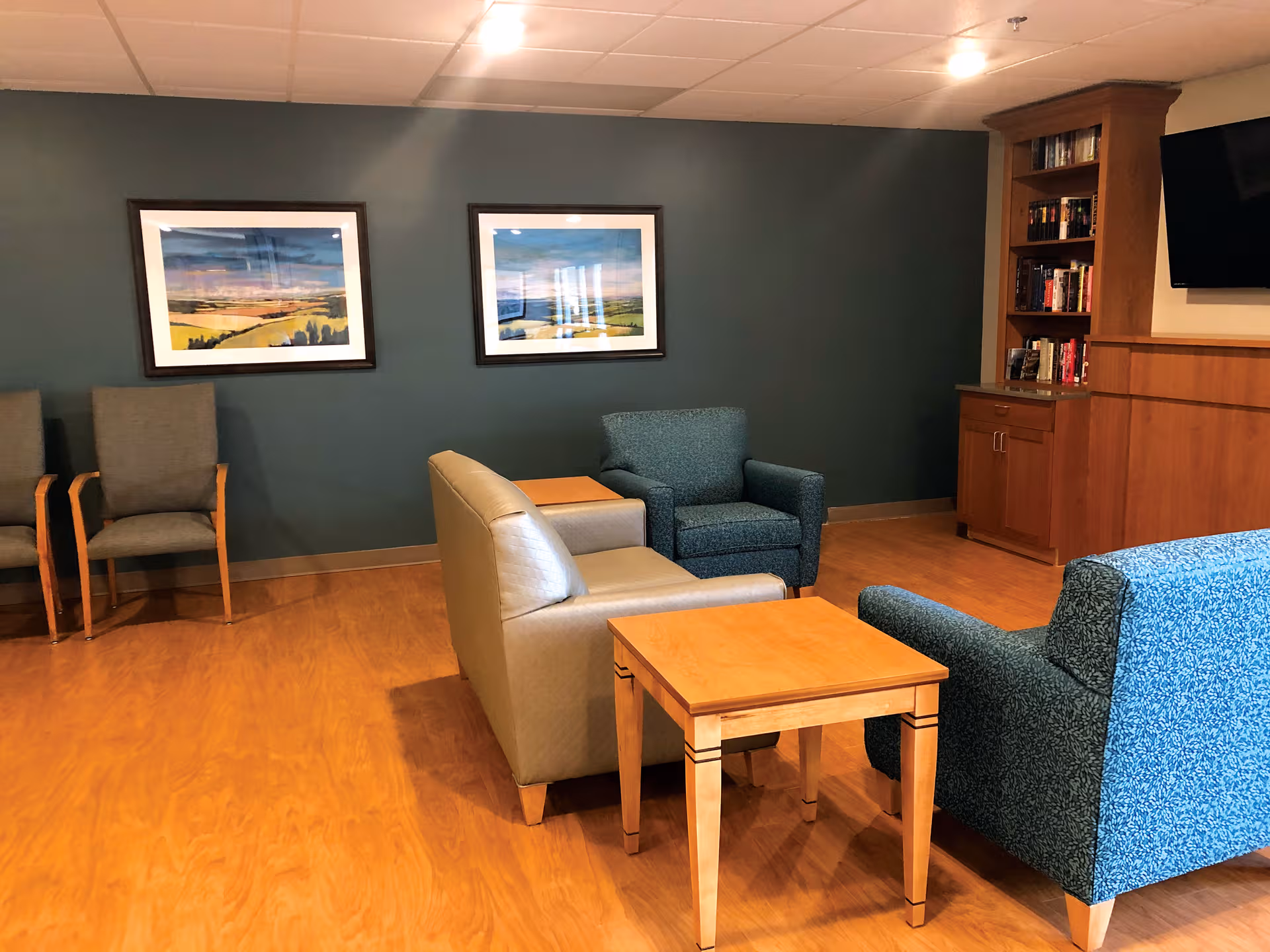 Senior living lounge with teal walls, framed landscape art, and assorted seating.