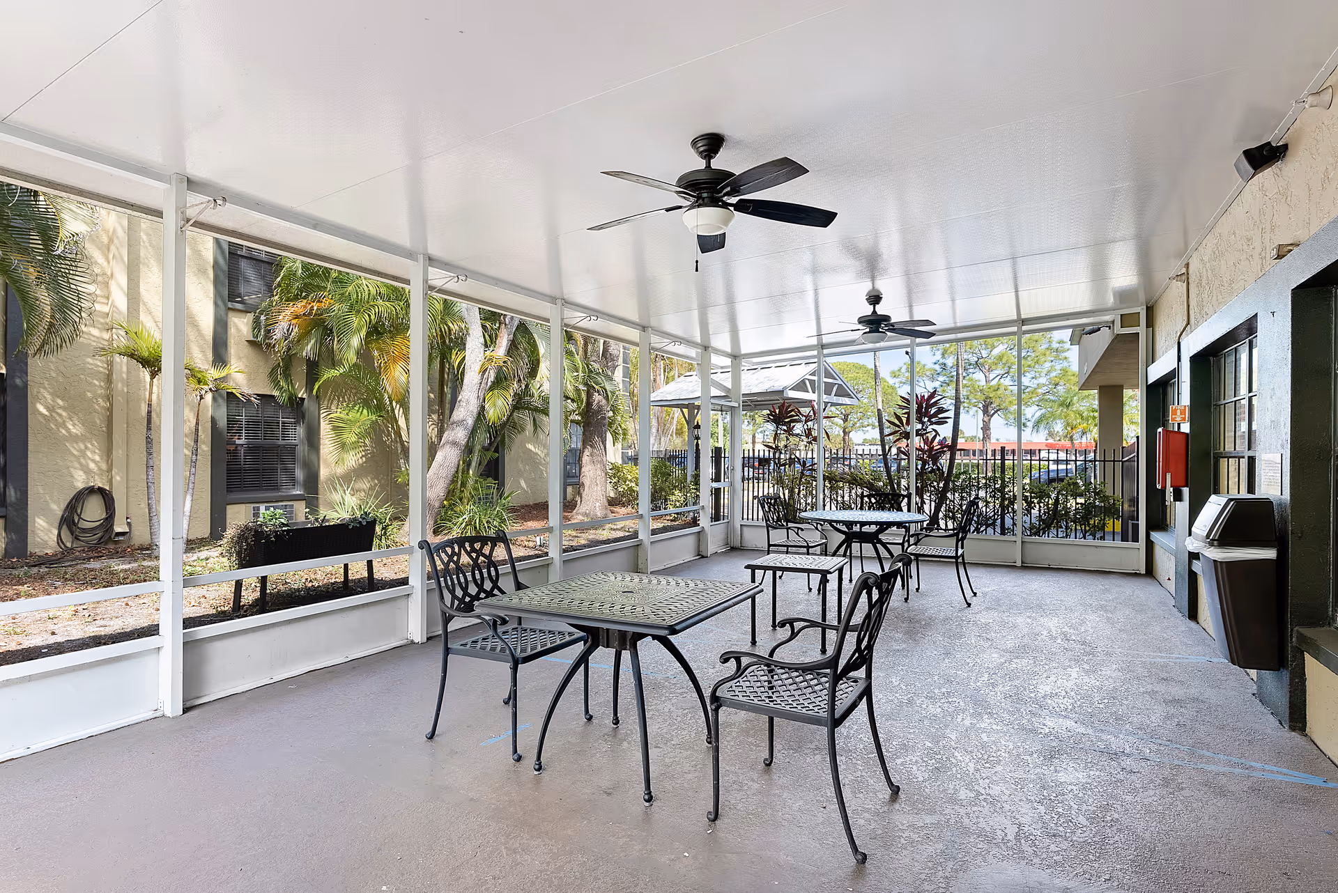 Covered outdoor patio area with metal tables and chairs, ceiling fans, and large screened windows overlooking a garden with palm trees and other plants.