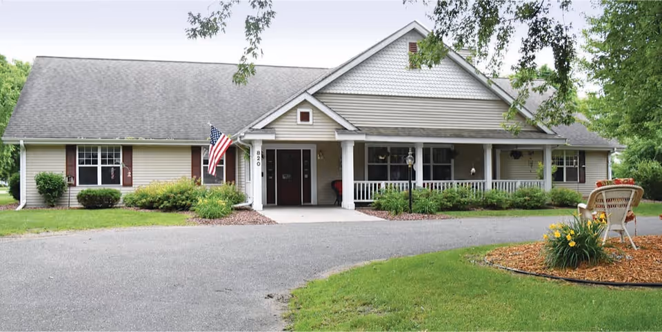 Single-story beige senior living building with a covered front porch, American flag, driveway and lawn.