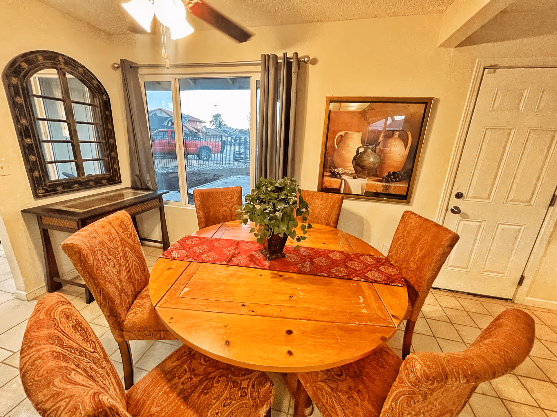 A dining area with a round wooden table covered partially by a red patterned table runner and a potted green plant centerpiece. Six upholstered chairs with a paisley pattern surround the table. Behind the table is a window with gray curtains, a decorative mirror on the left wall, and a framed painting of pottery on the right wall. A white door is visible on the right side of the image.