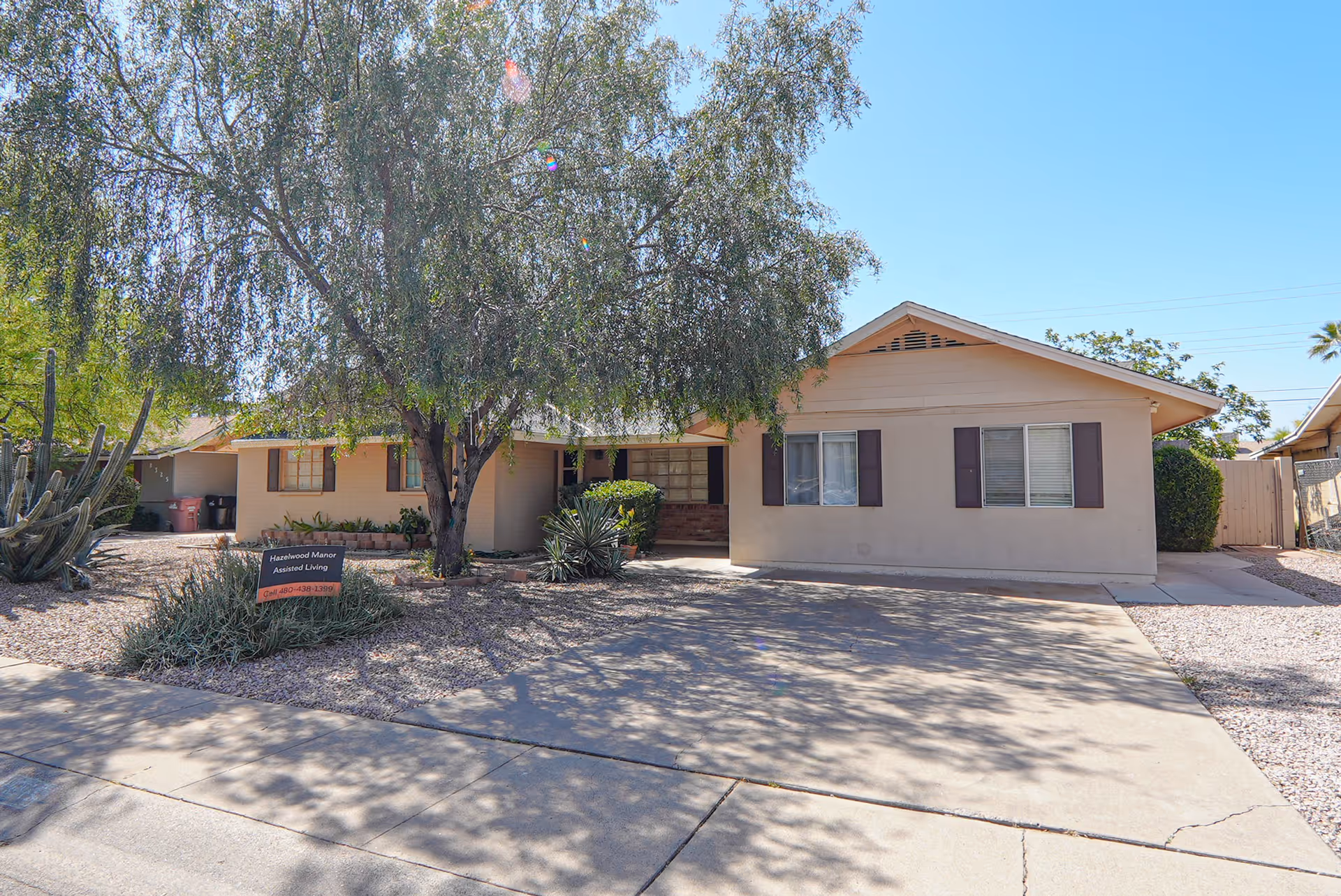 Exterior view of a single-story beige building with brown shutters, surrounded by desert landscaping including a large tree and cacti. A sign in front reads 'Hazelwood Manor Assisted Living'. The driveway and sidewalk are visible under a clear blue sky.