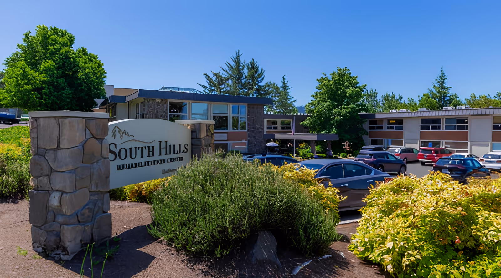 Entrance sign and front exterior of South Hills Rehabilitation Center with parked cars, landscaped bushes and trees under a clear blue sky.