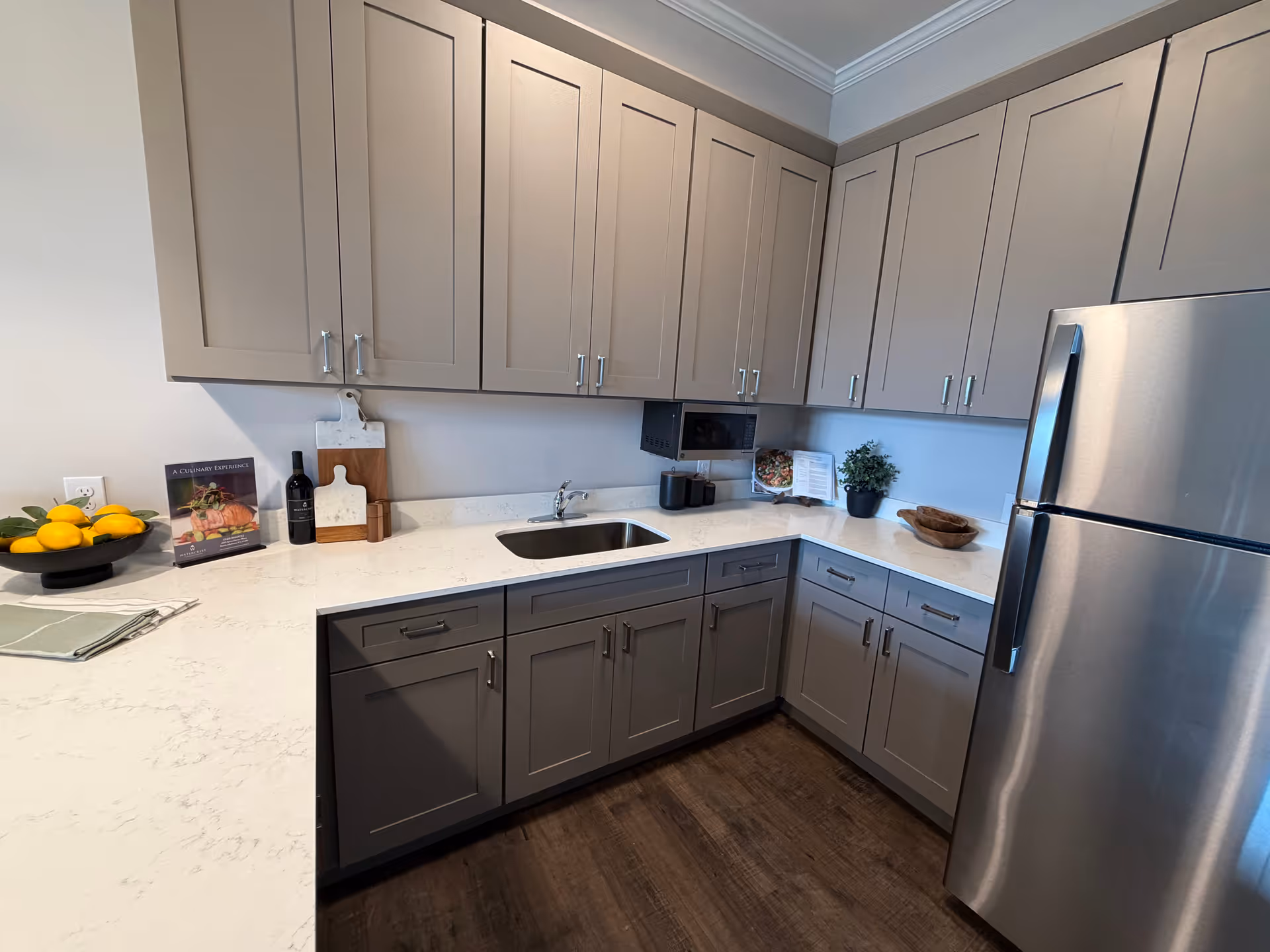 Modern L-shaped kitchen with gray cabinets, white countertops, sink, and stainless steel refrigerator.