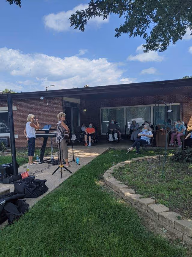 Outdoor scene at Prestige Care Center of Nebraska City showing a small musical performance with two women playing instruments and singing. Several elderly people are seated outside the building, some in wheelchairs, watching the performance. The sky is partly cloudy and there is a grassy area with a stone-bordered garden bed in the foreground.