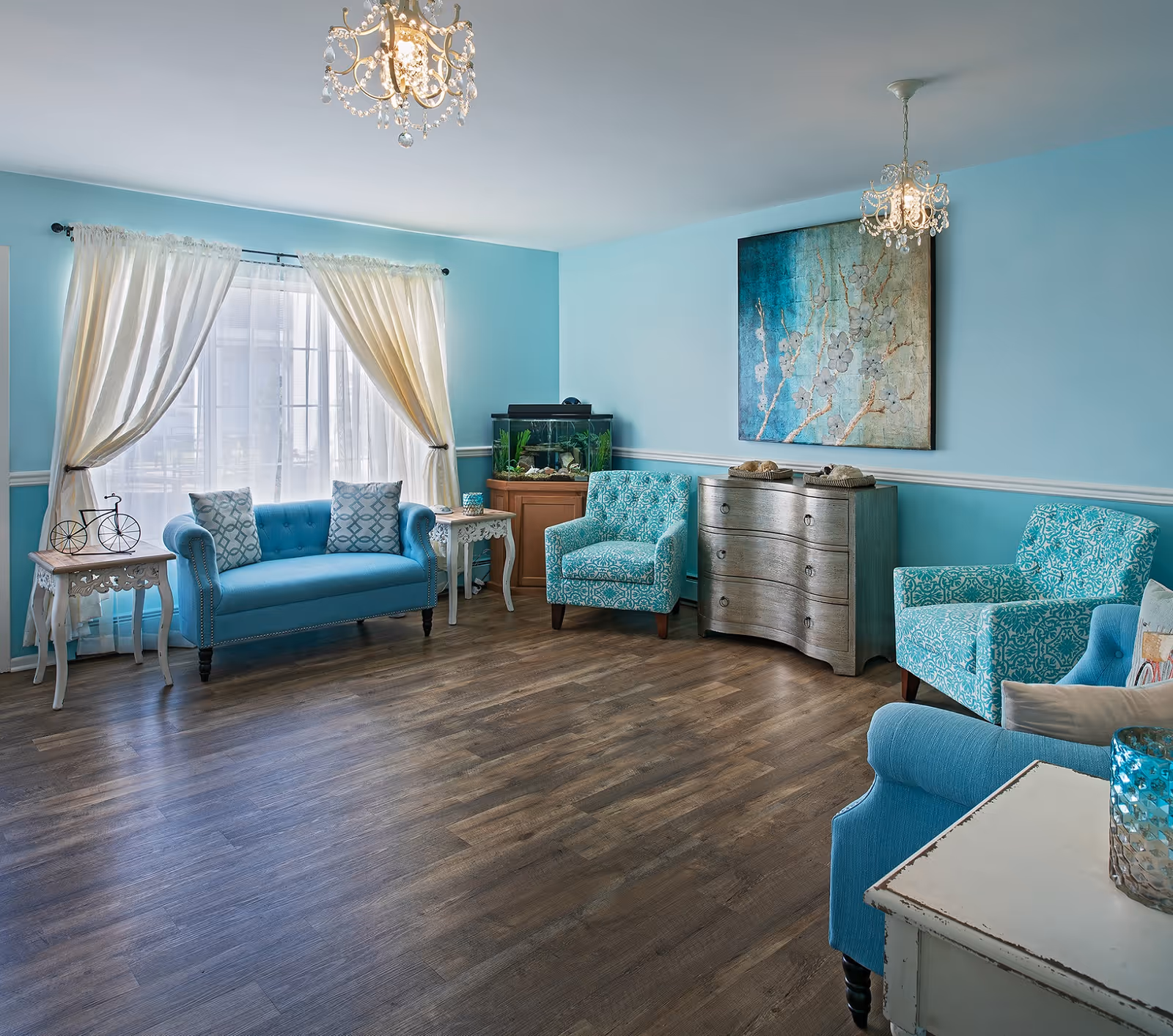 Bright turquoise living room with blue furniture, chandeliers, and large windows in a senior living facility.