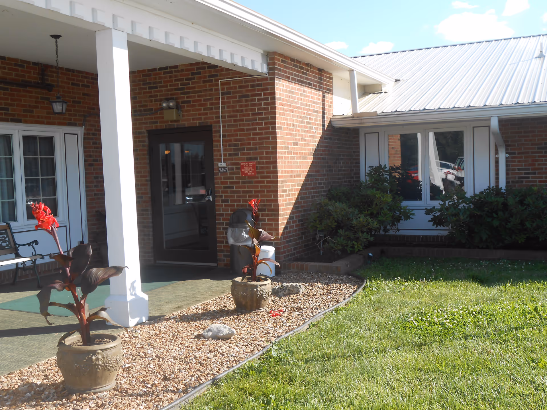 Exterior view of a brick building with a covered entrance supported by white columns. There are two potted plants with red flowers on a gravel bed next to a green lawn. A bench is visible near a window on the left side, and a glass door leads inside the building.