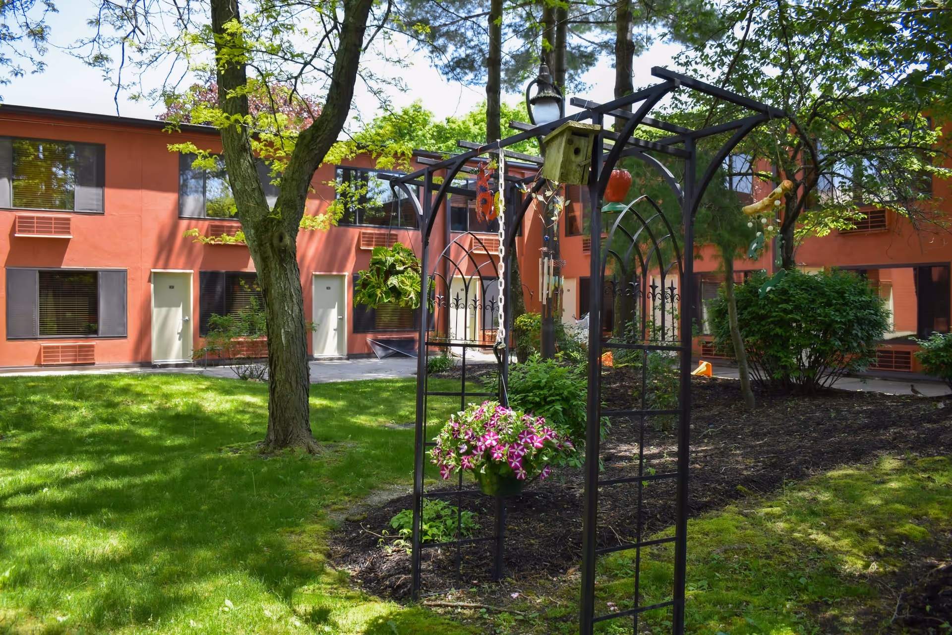 A courtyard garden area with green grass, trees, and shrubs in front of a two-story building with orange walls and multiple windows and doors. A black metal garden arch with hanging flower pots and a birdhouse is in the foreground.