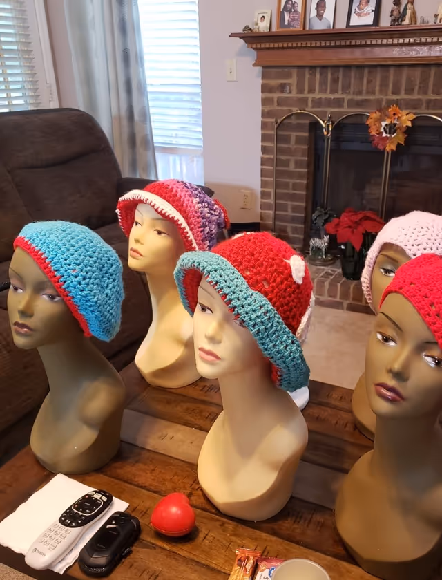 Mannequin heads wearing crocheted hats arranged on a wooden table near a brick fireplace.