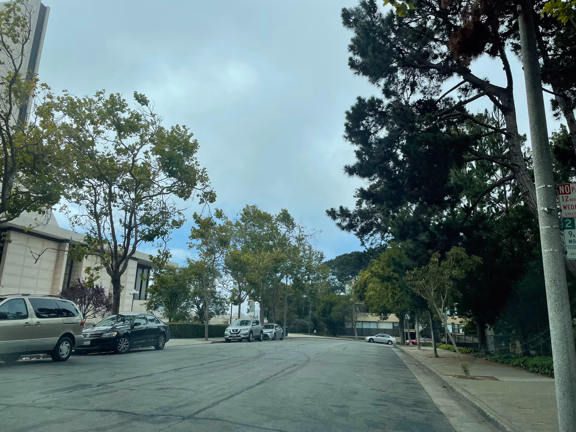 A quiet residential street with parked cars, trees, and a sidewalk along a building.