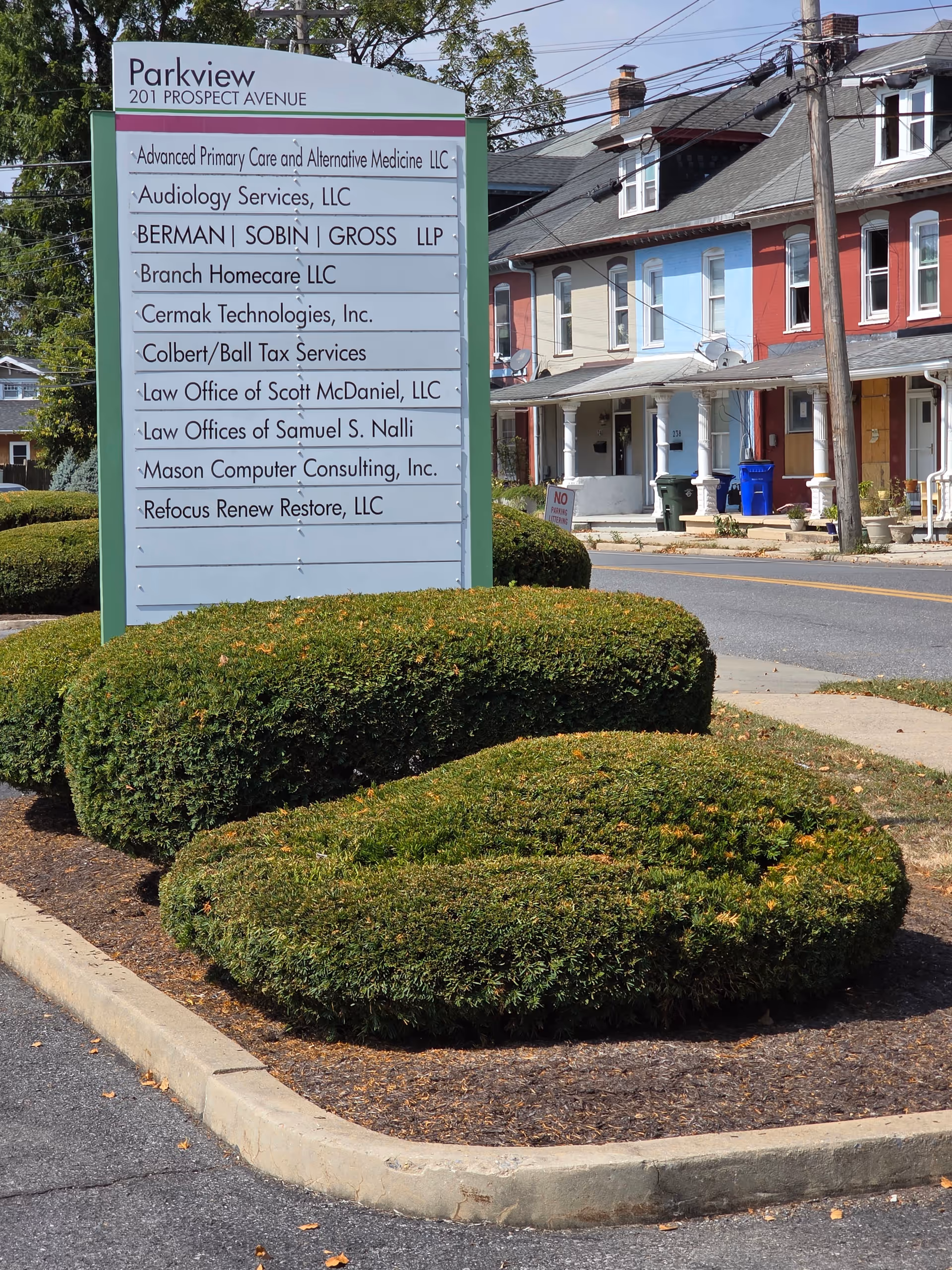 Large exterior directory sign listing tenants, with trimmed shrubs and row of houses nearby.