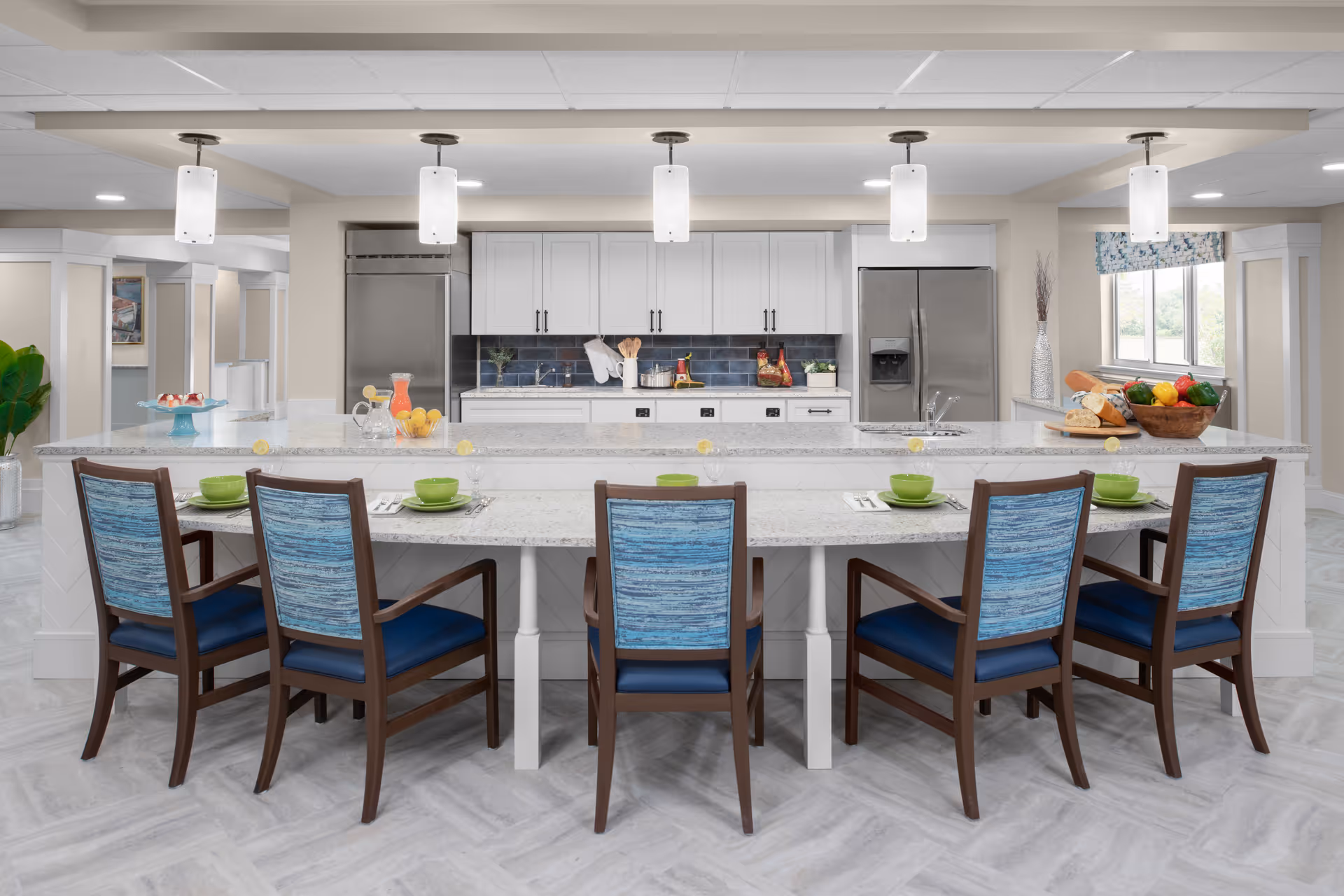 A modern kitchen and dining area with a long white marble island countertop surrounded by six wooden chairs with blue cushions and backs. The kitchen features white cabinets, stainless steel refrigerator and freezer, a sink, and a blue tile backsplash. The countertop is set with green bowls, plates, and glasses, and there are decorative items including a bowl of fruit, a pitcher of juice, and a cake stand with desserts. Four pendant lights hang from the ceiling above the island.