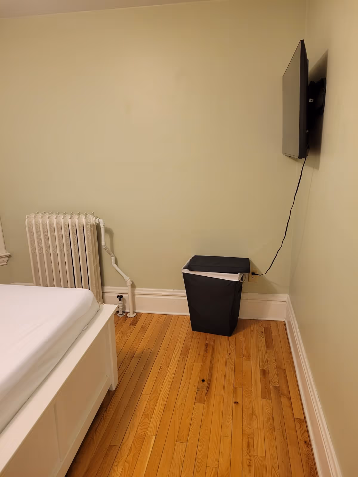 Small bedroom in a senior living facility with bed, radiator, trash can, and wall TV.
