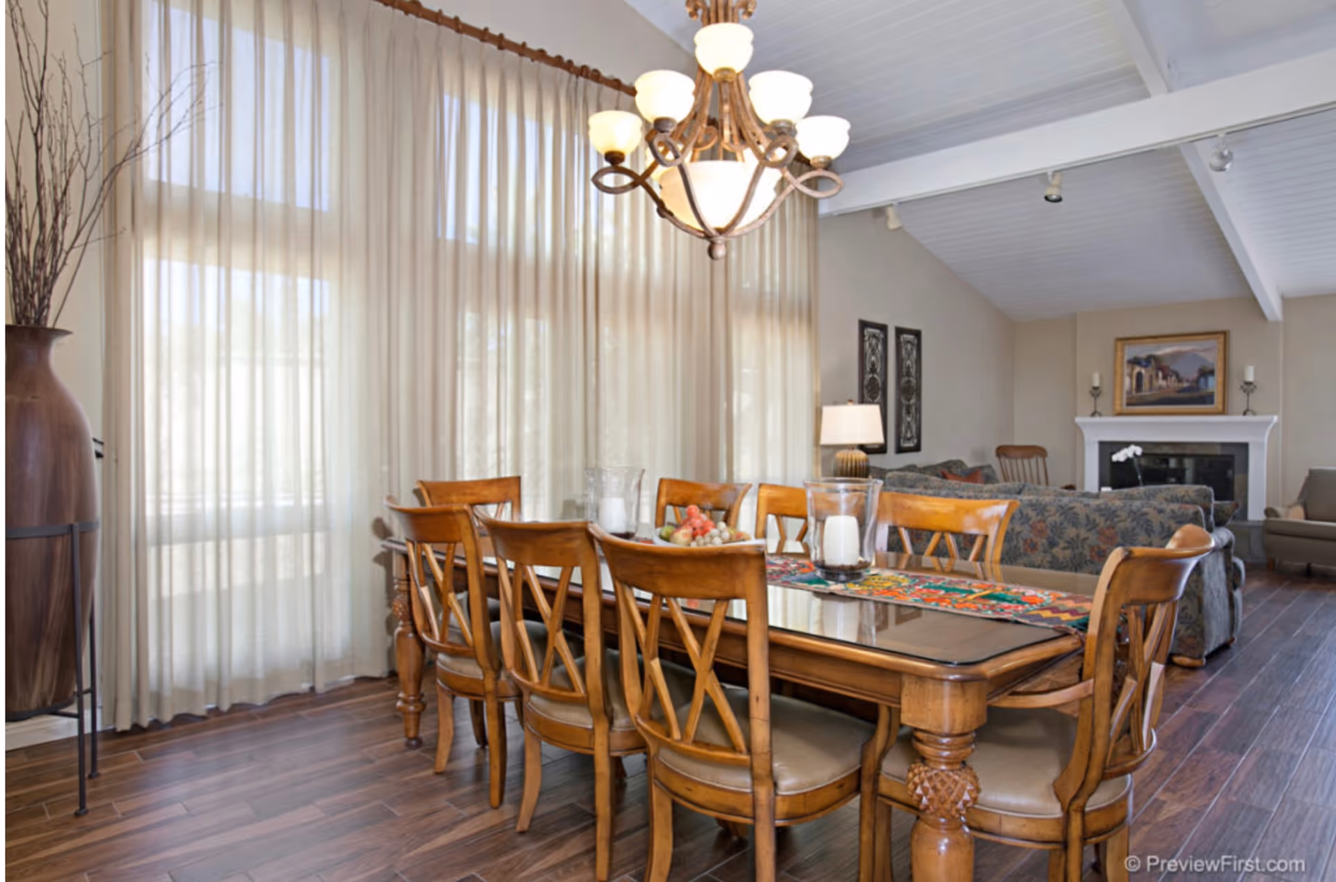 A spacious dining room with a large wooden table, chairs, a chandelier, and sheer curtains.