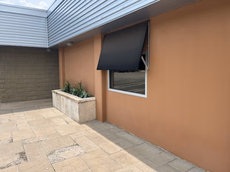 Exterior view of a peach-colored building with a window, black awning, and planter.