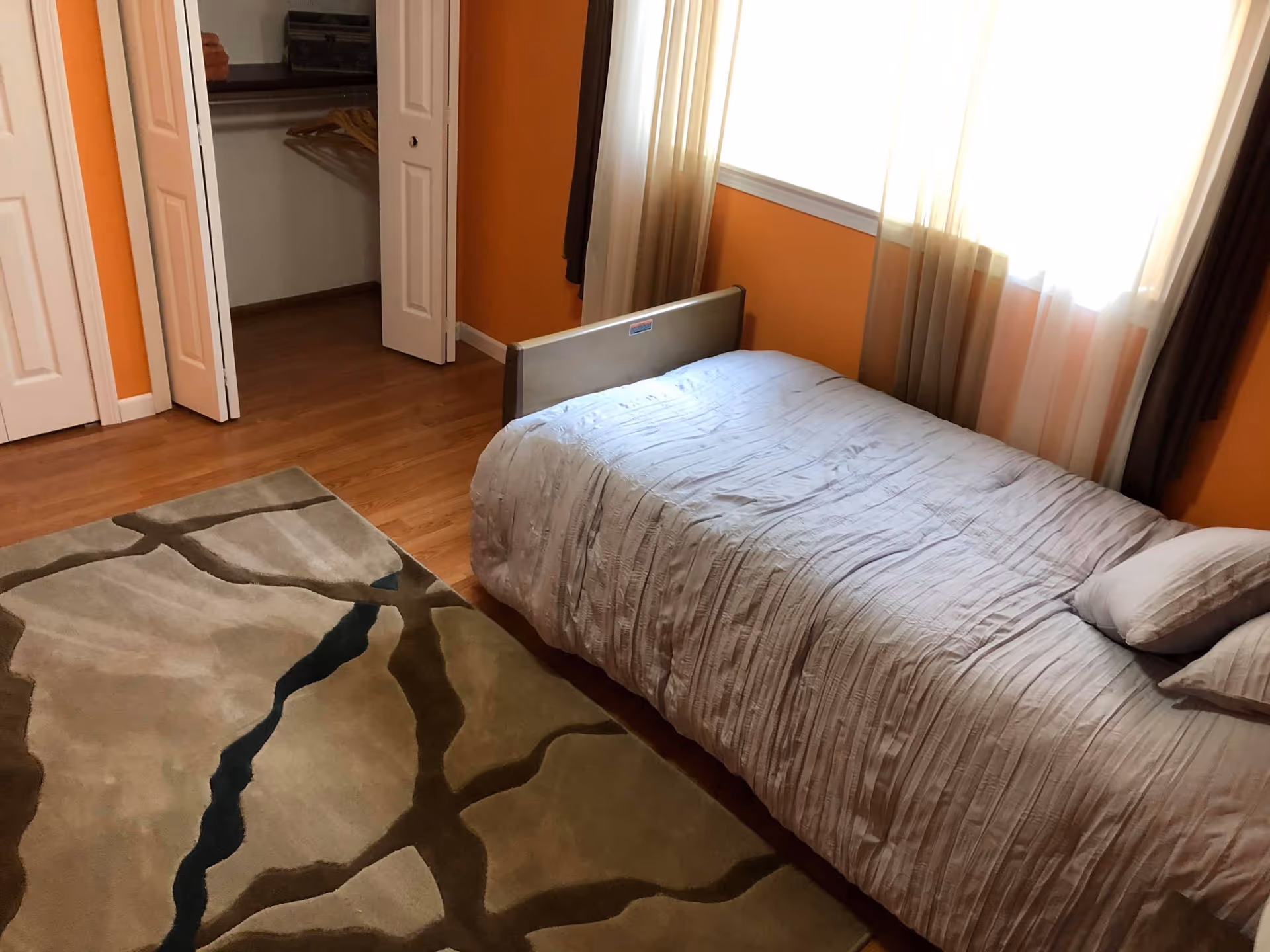 A bedroom with orange walls and wooden flooring featuring a single bed with a light gray bedspread and two pillows. There is a patterned area rug on the floor and a window with sheer curtains letting in natural light. An open closet with hangers is visible in the corner.
