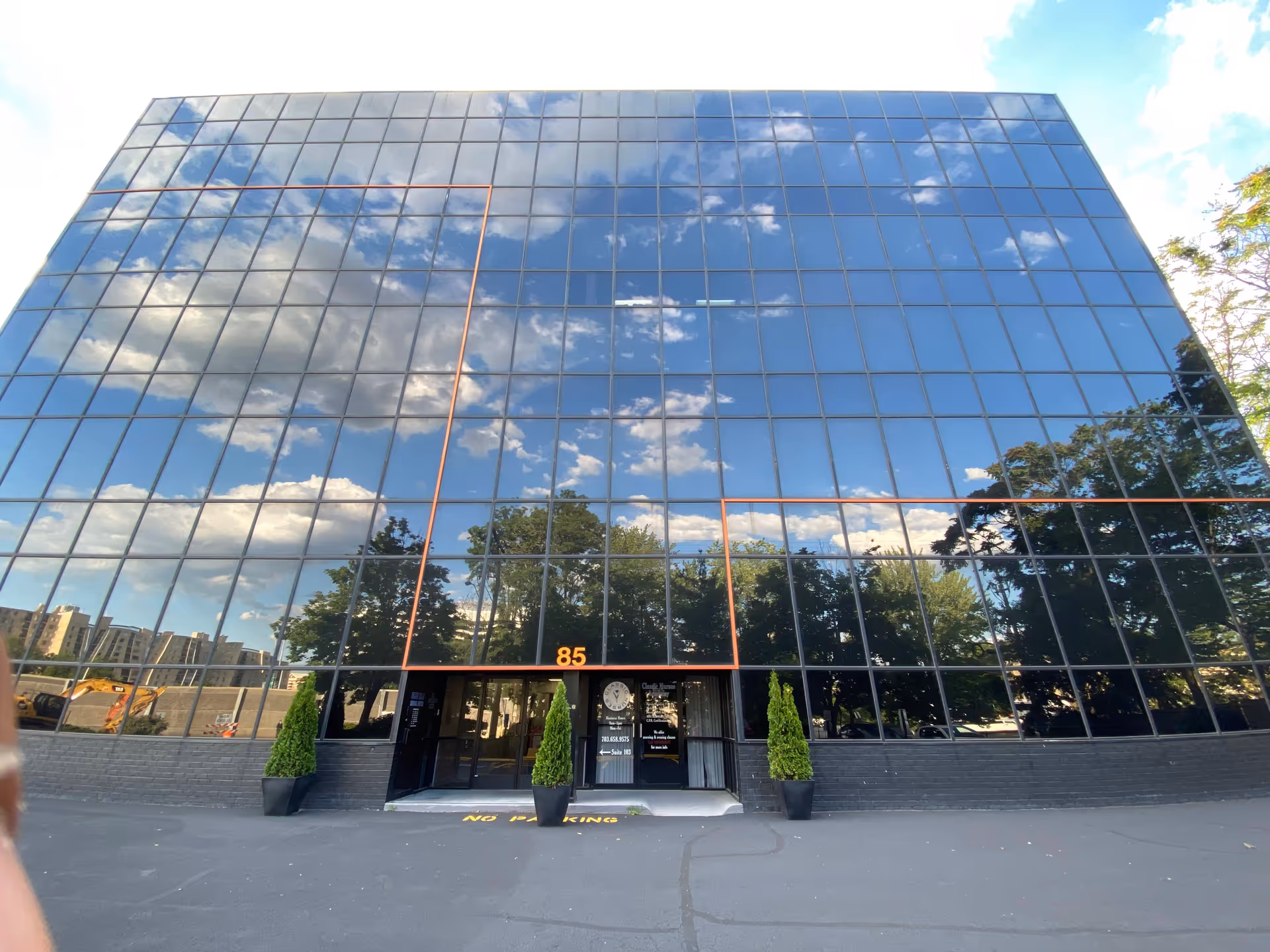 A tall glass-front office building with reflective windows and an entrance flanked by potted trees.
