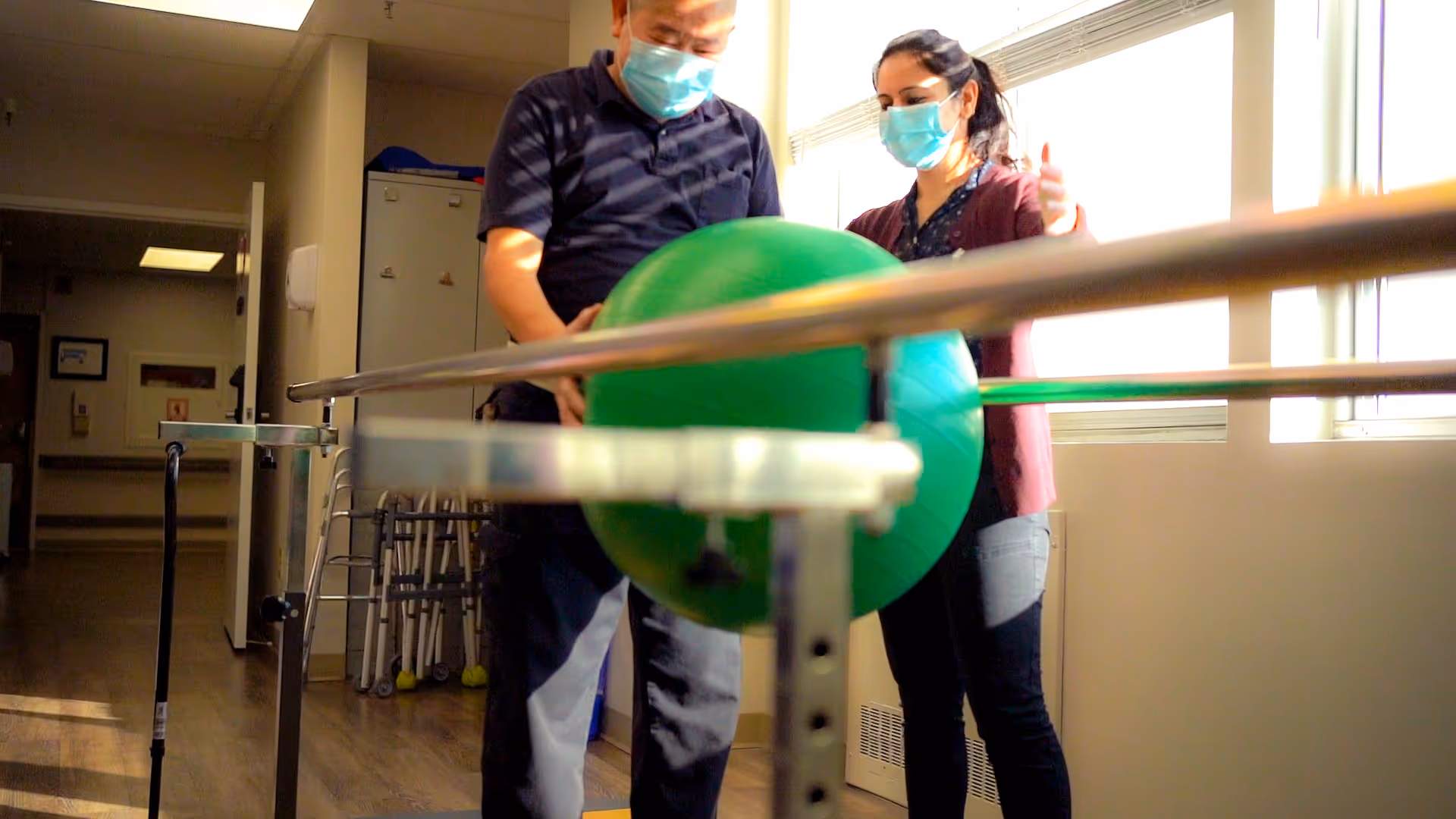 A man and a woman wearing face masks are engaged in a physical therapy exercise using a large green exercise ball and parallel bars in a well-lit room with wooden flooring and medical equipment in the background.
