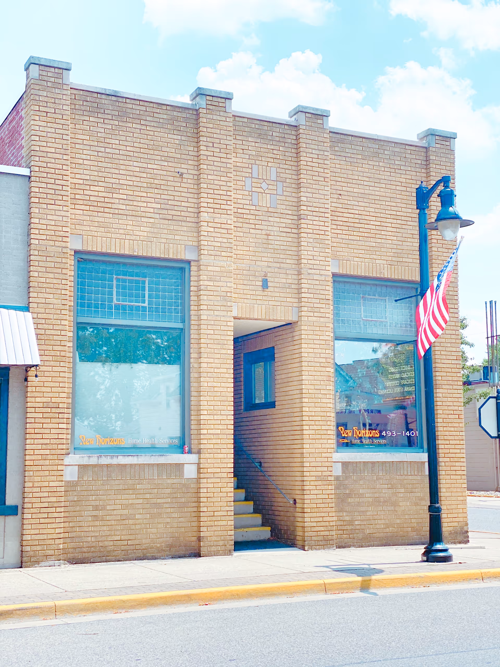 Brick storefront with large windows, entry stairs, and a waving American flag.