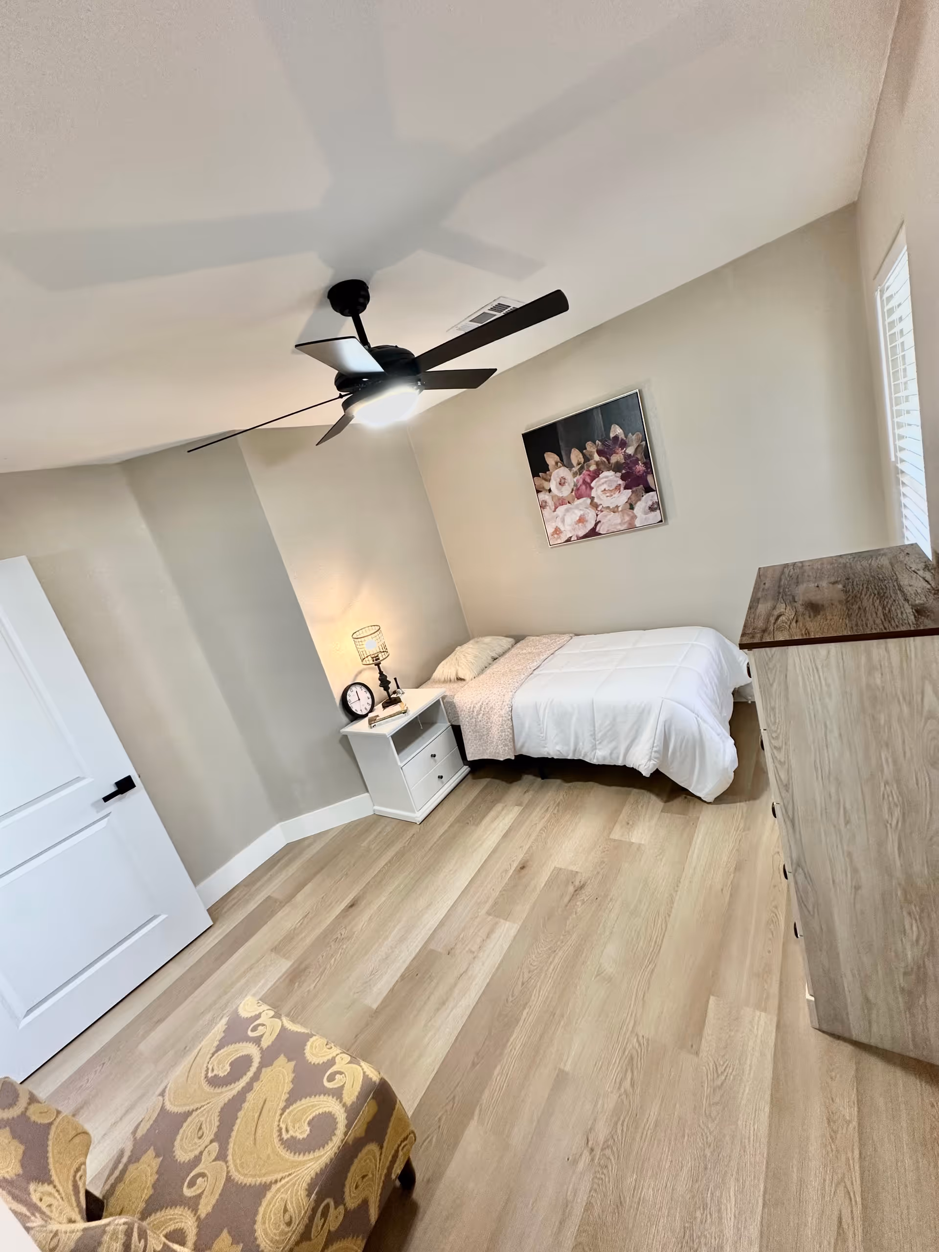 Cozy bedroom with bed, nightstand, dresser, ceiling fan, and floral wall art.