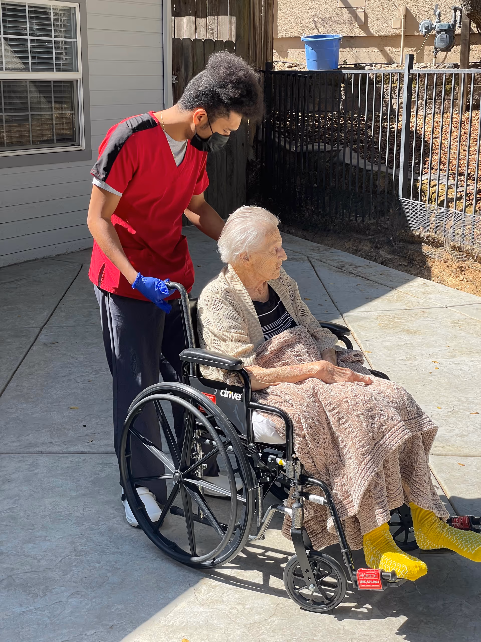 A caregiver wearing a red scrub top, black face mask, and blue gloves is pushing an elderly woman in a wheelchair outdoors on a paved area. The elderly woman is covered with a knitted blanket and wearing bright yellow socks. They are near a building with a window and a wooden fence.