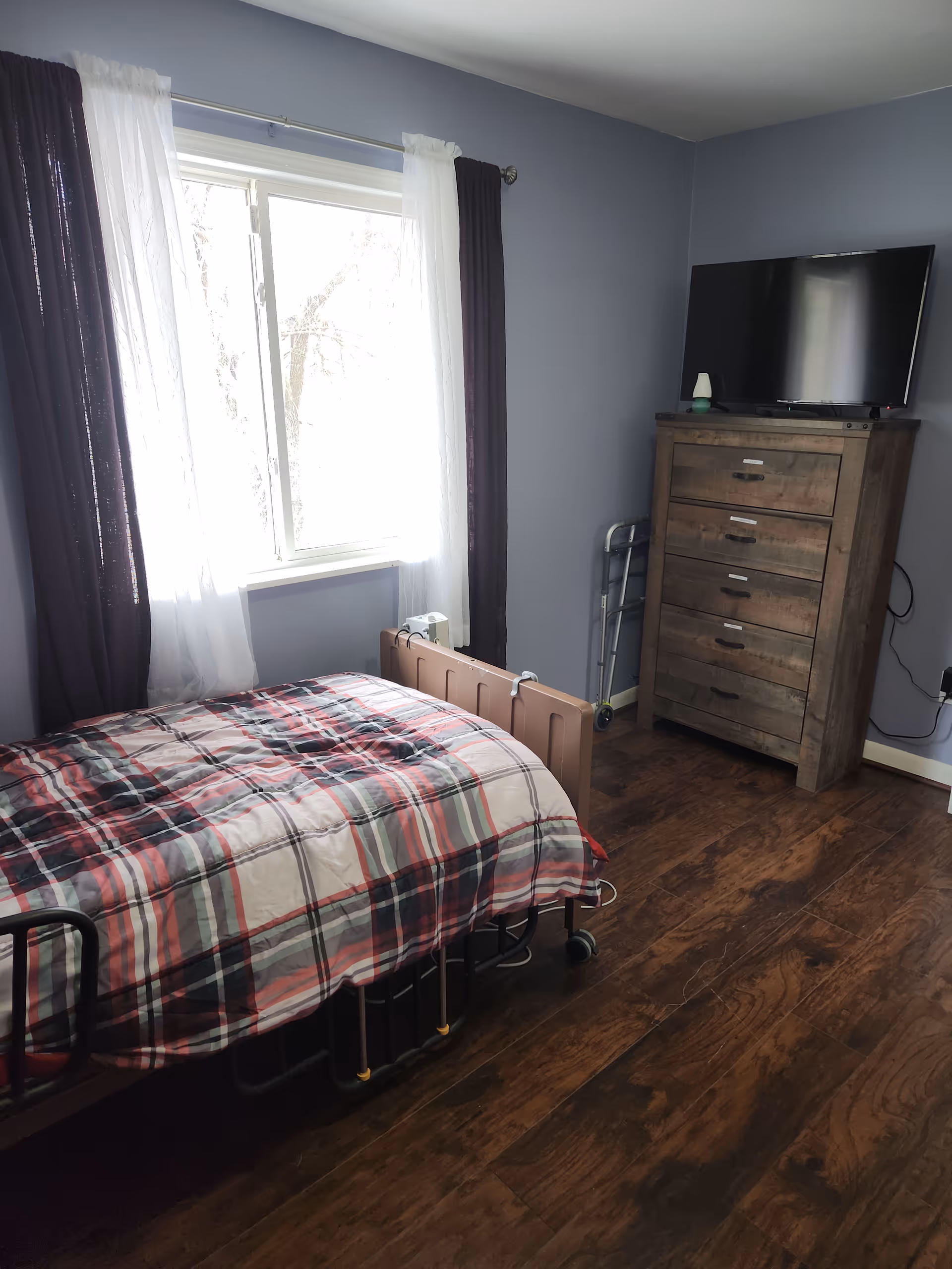 A furnished bedroom with a plaid bed, dresser with TV, walker, and window.