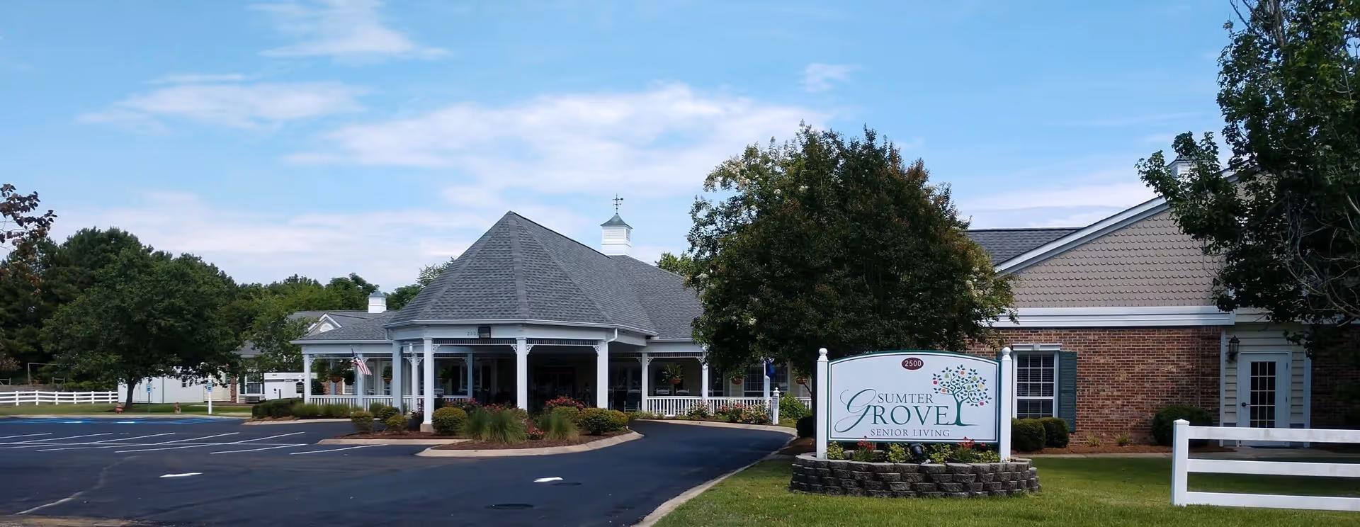 Exterior view of Sumter Grove Senior Living facility showing a large building with a covered entrance, surrounded by trees and a well-maintained lawn. There is a sign in front with the facility's name and a paved driveway leading to the entrance.