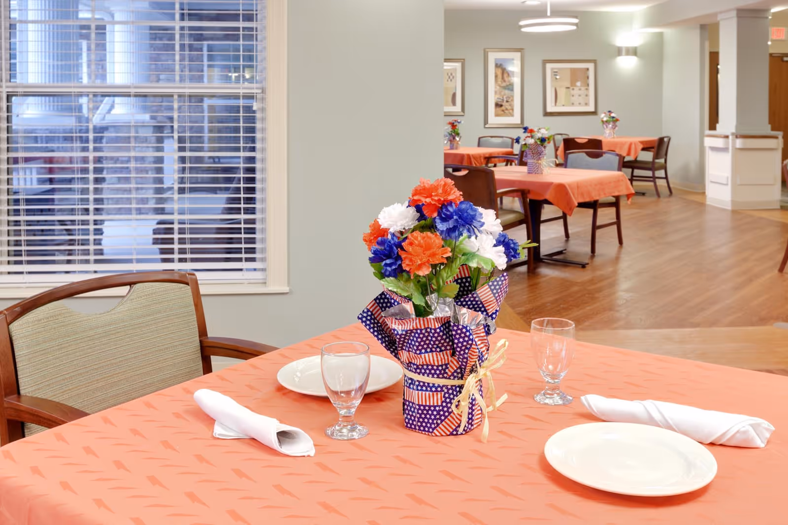 Senior living facility dining room with orange tablecloths, flower centerpieces, and framed wall art.